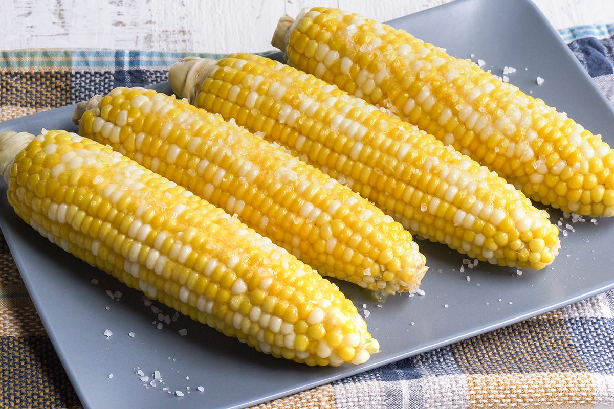Four ears of corn on the cob, lightly buttered and sprinkled with salt, are arranged on a gray plate set on a plaid cloth.