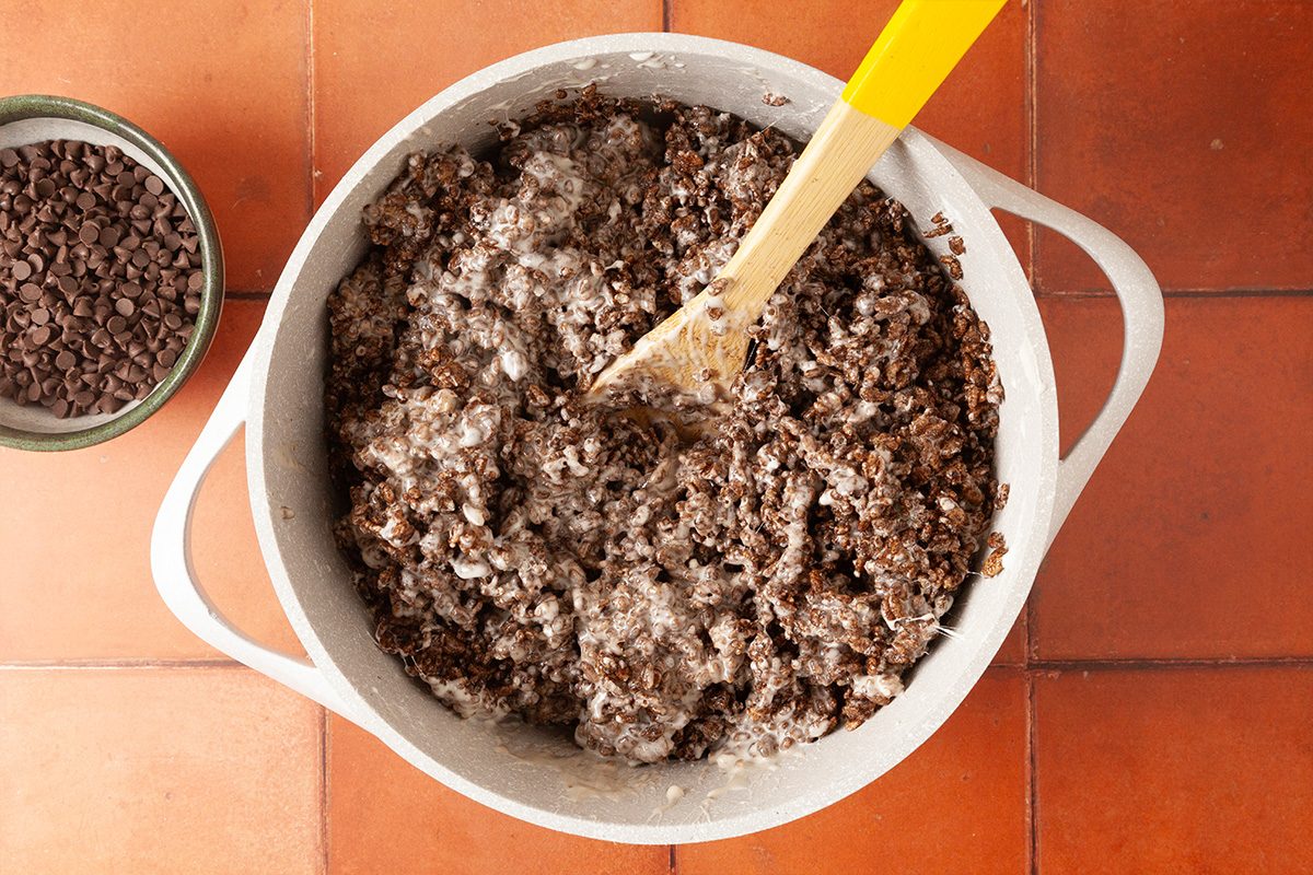A white pot filled with a chunky mixture being stirred by a yellow spatula, sitting on a tiled surface. A small bowl of chocolate chips is placed next to the pot.