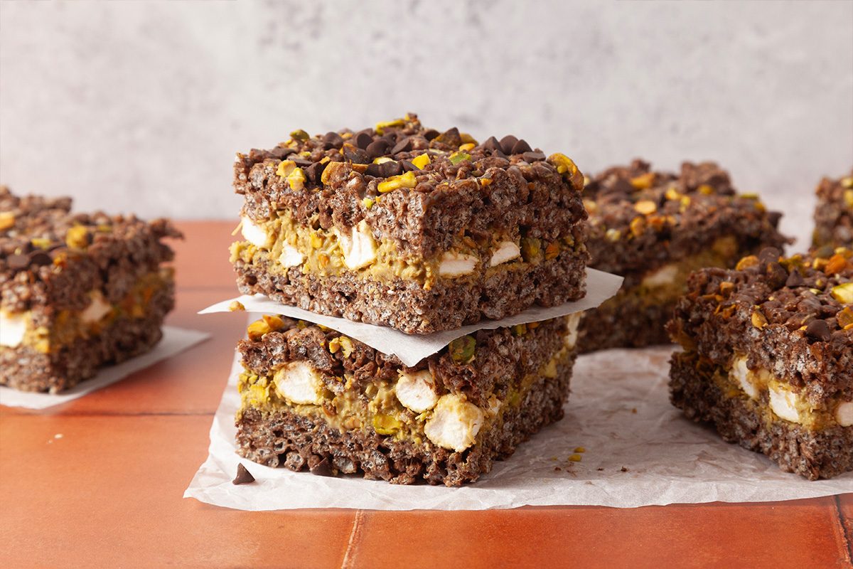 Four chocolate crumb bars with marshmallows and a crumbly topping are stacked and arranged on a sheet of parchment paper, set against a neutral background on a terracotta surface.