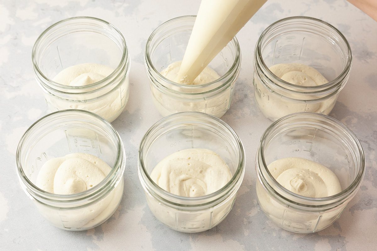 Six glass jars are arranged on a light surface, each filled with a swirl of creamy white mixture. A piping bag is shown dispensing more of the mixture into one of the jars.