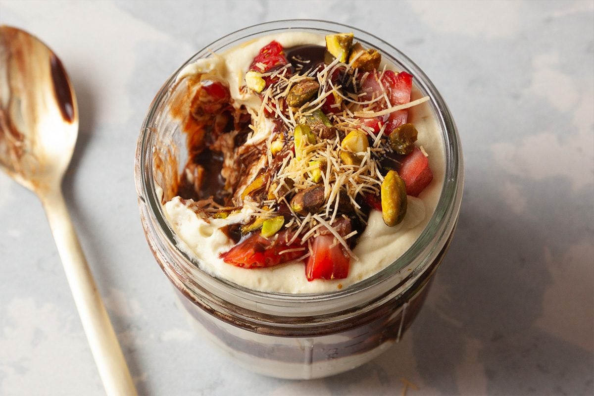 A glass jar filled with yogurt, chocolate sauce, granola, chopped strawberries, shredded coconut, and pistachios. A spoon with a chocolate smear rests beside the jar on a light surface.A glass jar filled with yogurt, chocolate sauce, granola, chopped strawberries, shredded coconut, and pistachios. A spoon with a chocolate smear rests beside the jar on a light surface.