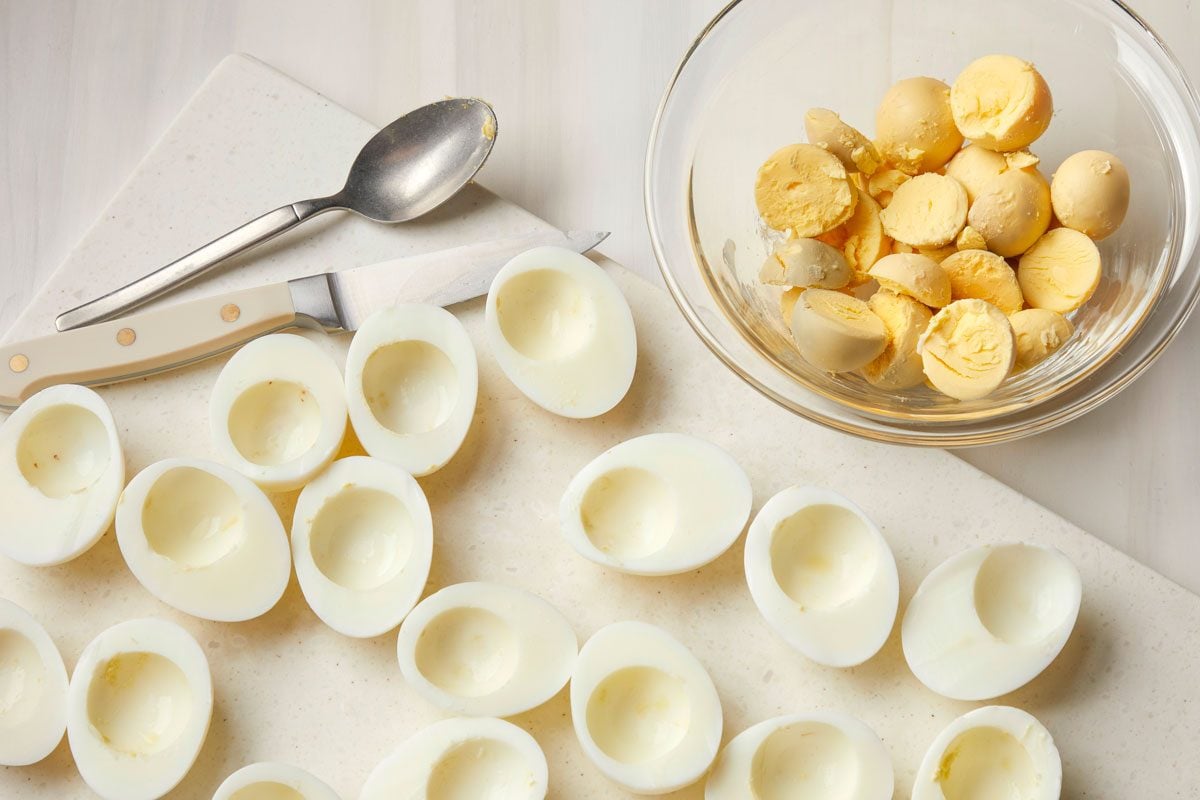 Overhead shot of eggs cut in half lengthwise on a cutting board with a spoon and a knife; Remove the yolks and save them in a glass bowl, reserving the whites; All set on a cream-colored surface;