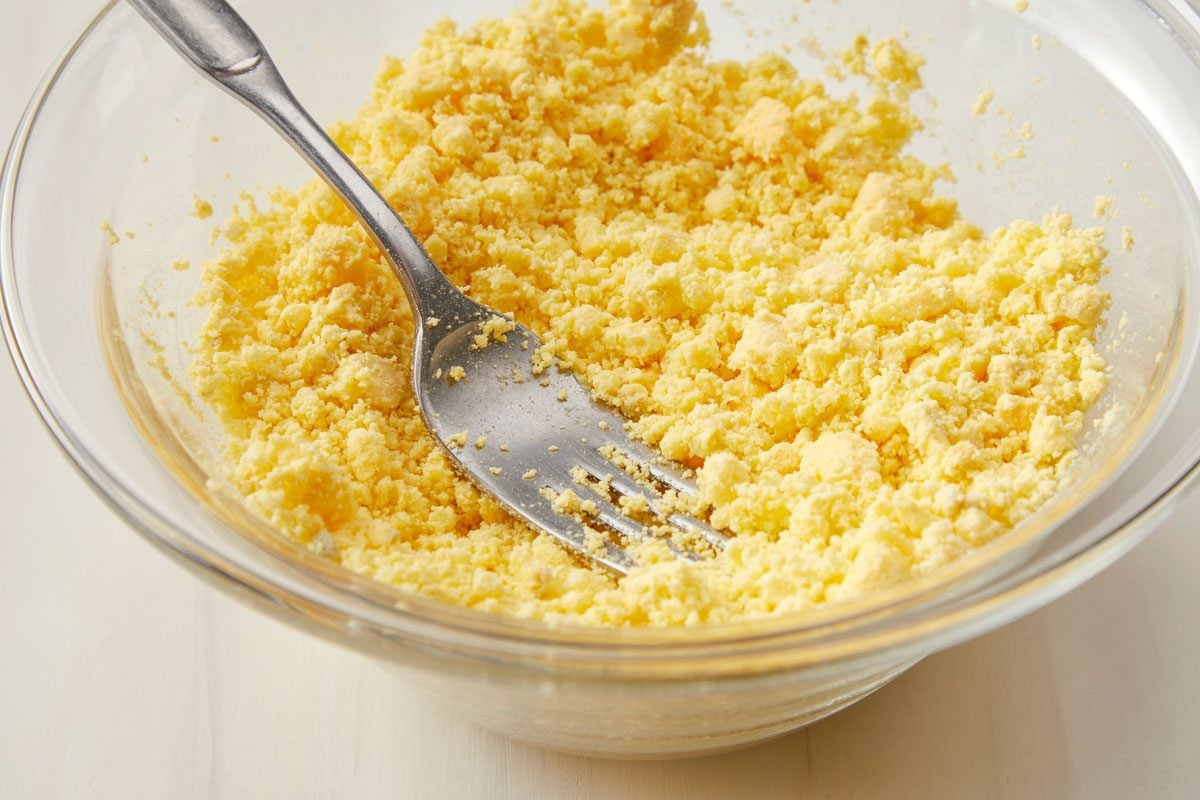 Closeup shot of a small glass bowl filled yolks; mash yolks with a metal fork; all set on a cream-colored surface;
