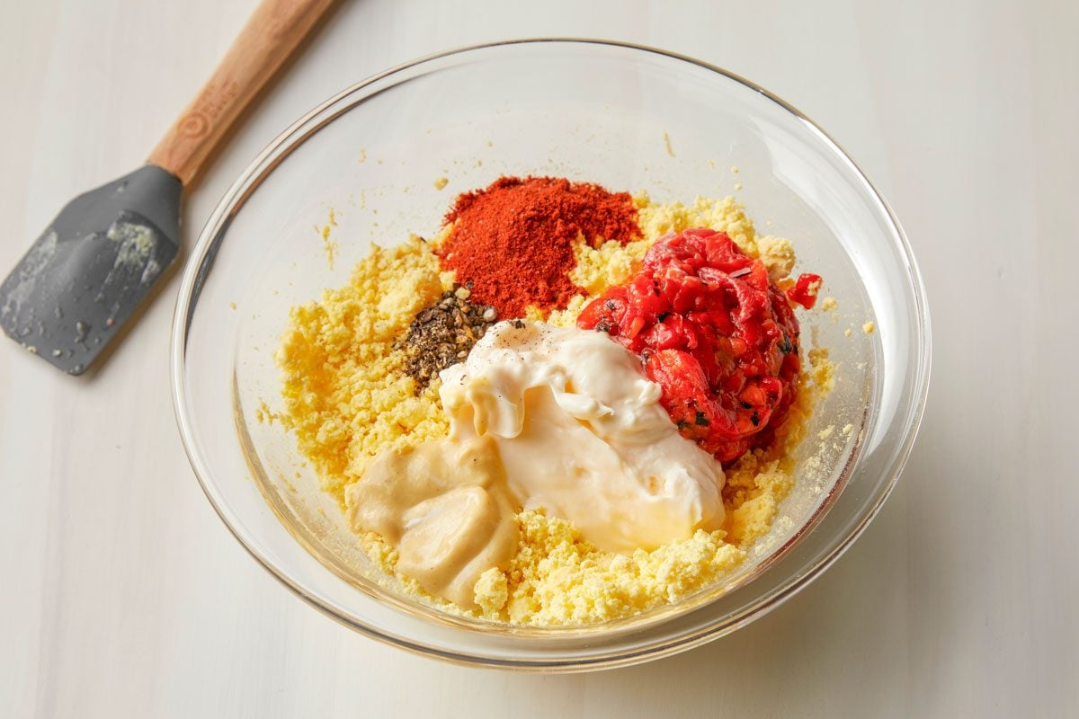 3/4 angle view shot of a glass bowl containing egg yolks; stir in mayonnaise; peppers; mustard; vinegar; paprika; salt and pepper until blended; spatula; all set on a cream-colored surface;