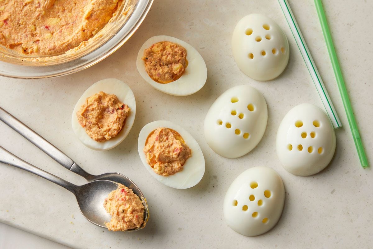Overhead shot of a small and a large straw; decorate each egg white to make a skull with eyes, a nose and a mouth; Spoon or pipe yolk mixture into egg whites; all set on marble board;