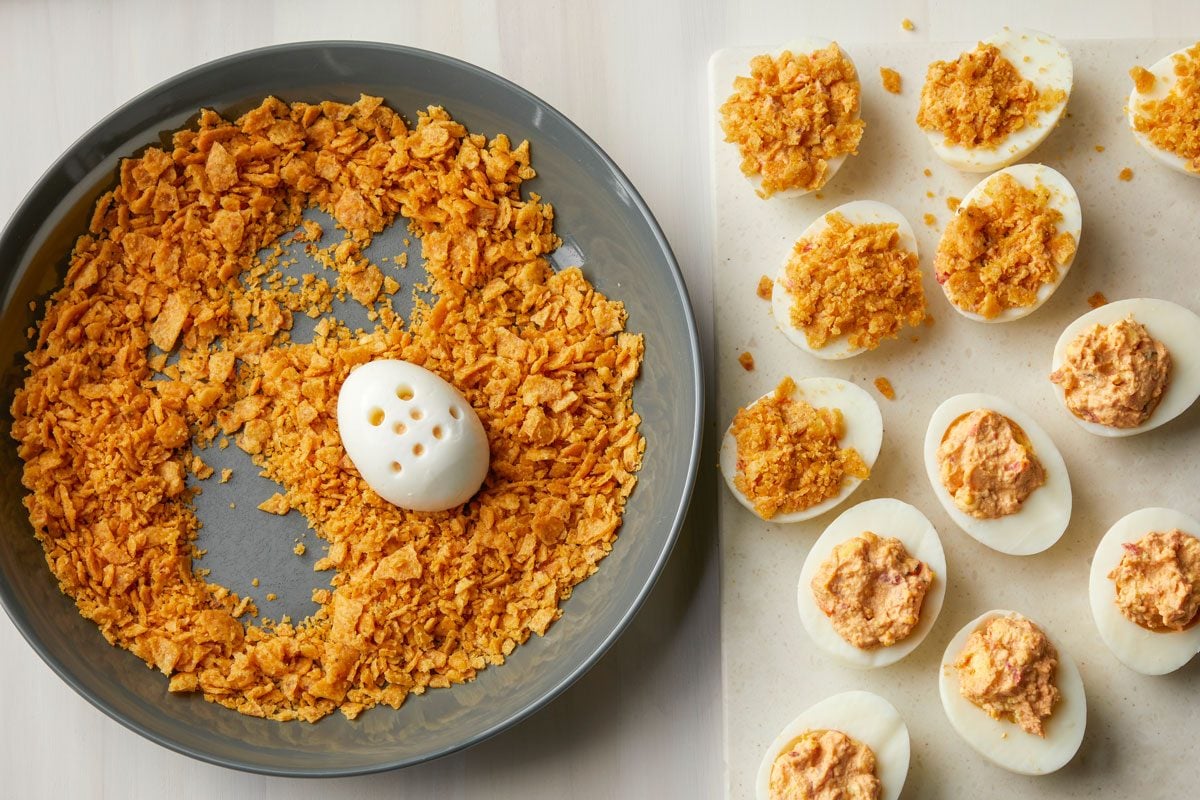 Overhead shot of place crushed chips in a shallow bowl; Dip each exposed yolk into chips; refrigerate covered until serving; all set on a cream-colored surface;