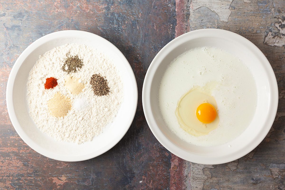 Two white bowls on a textured surface; one bowl contains flour, spices, and seasonings, while the other contains milk and a raw egg.