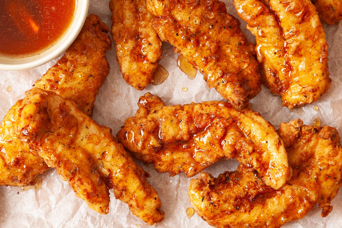 Crispy fried chicken tenders glazed with sauce, arranged on parchment paper with a small bowl of dipping sauce in the corner.