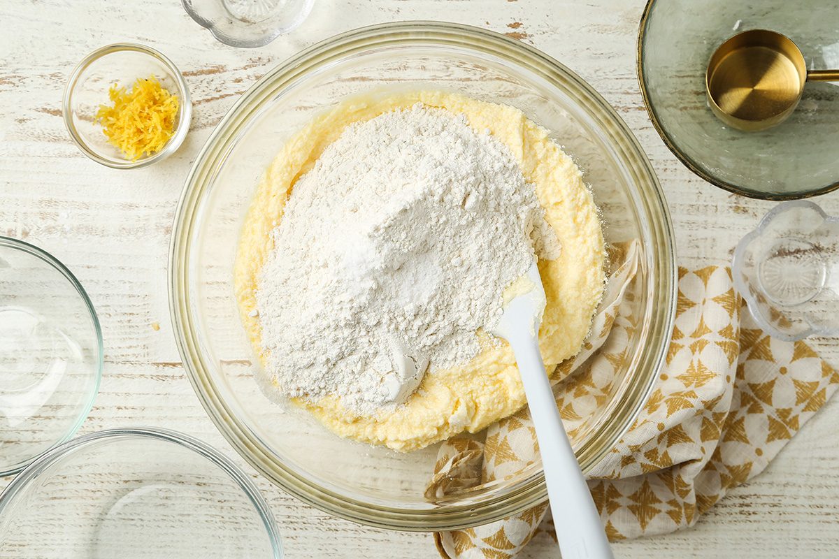 A mixing bowl with flour and creamed butter mixture being combined with a white spatula. Nearby are a small bowl of lemon zest, a glass with liquid, and an empty patterned kitchen towel.
