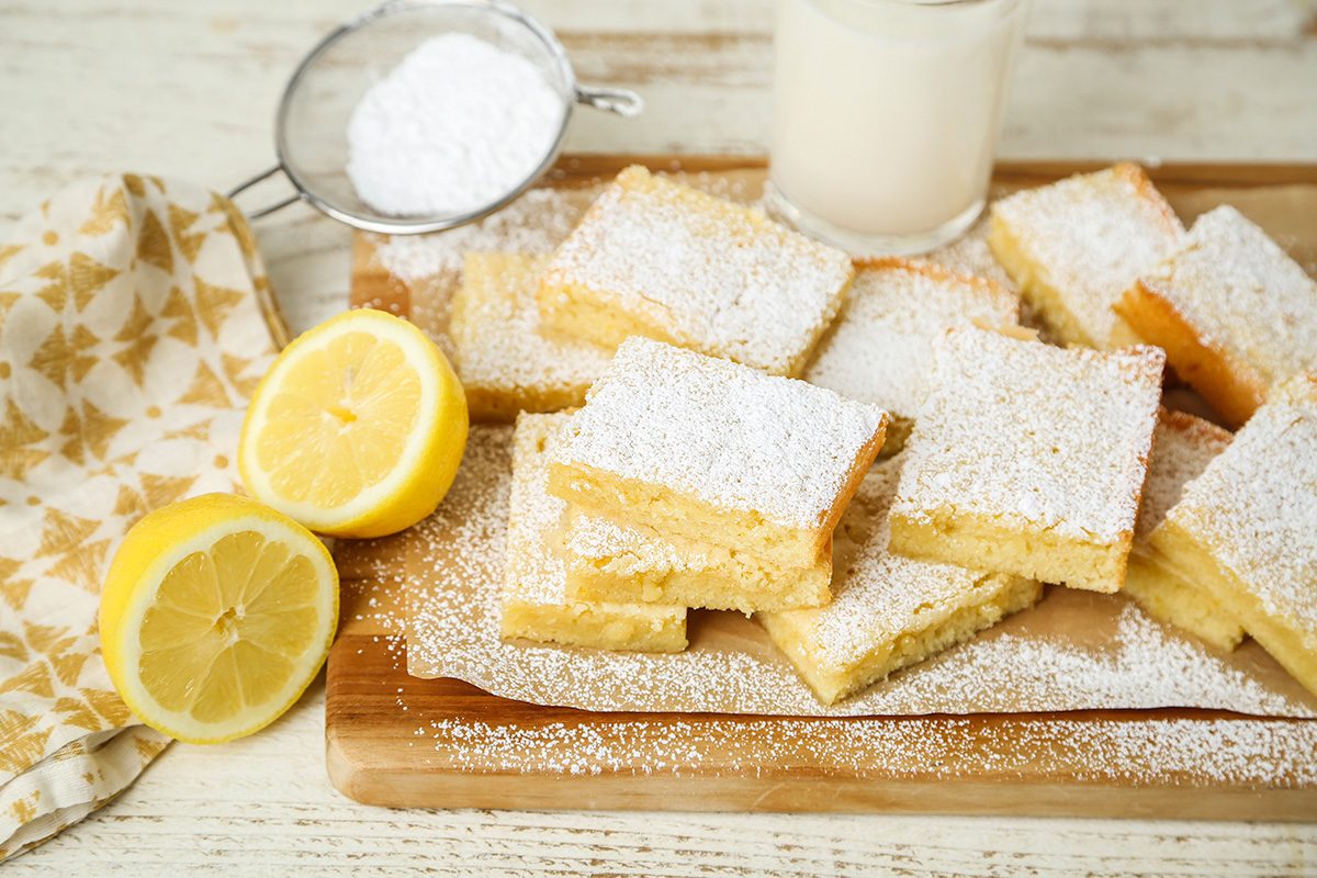 Lemon bars dusted with powdered sugar are stacked on a wooden board next to sliced lemons, a sifter with powdered sugar, a glass of milk, and a patterned napkin.