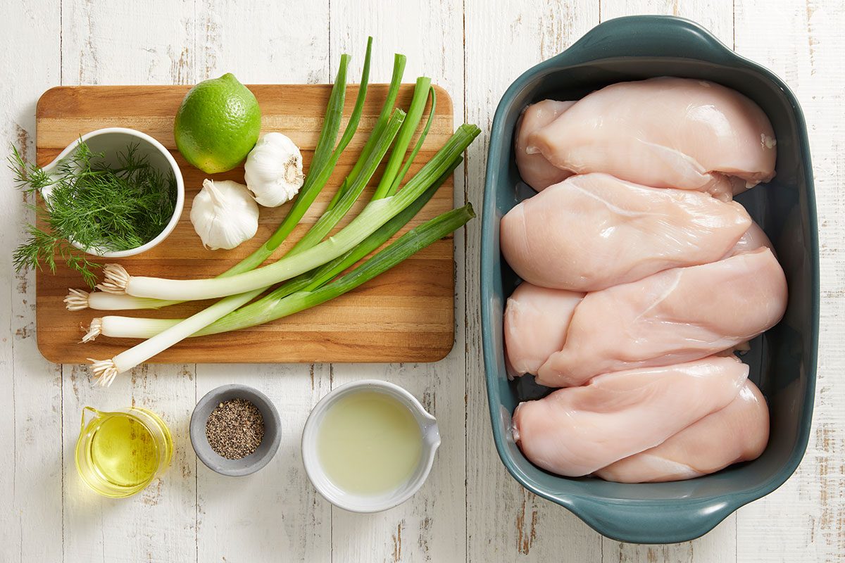 Raw chicken breasts in a baking dish, alongside green onions, garlic, a lime, fresh dill, olive oil, pepper, and lemon juice on a wooden cutting board on a white surface.