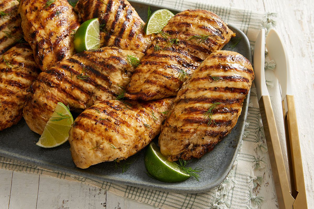 Grilled chicken breasts with dark grill marks, served on a gray plate with lime wedges, placed on a light plaid cloth and accompanied by a set of tongs on the side.