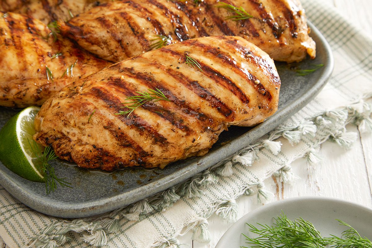 Grilled chicken breasts with charred grill marks are served on a gray plate, garnished with fresh dill and a lime wedge, placed on a light, textured cloth.