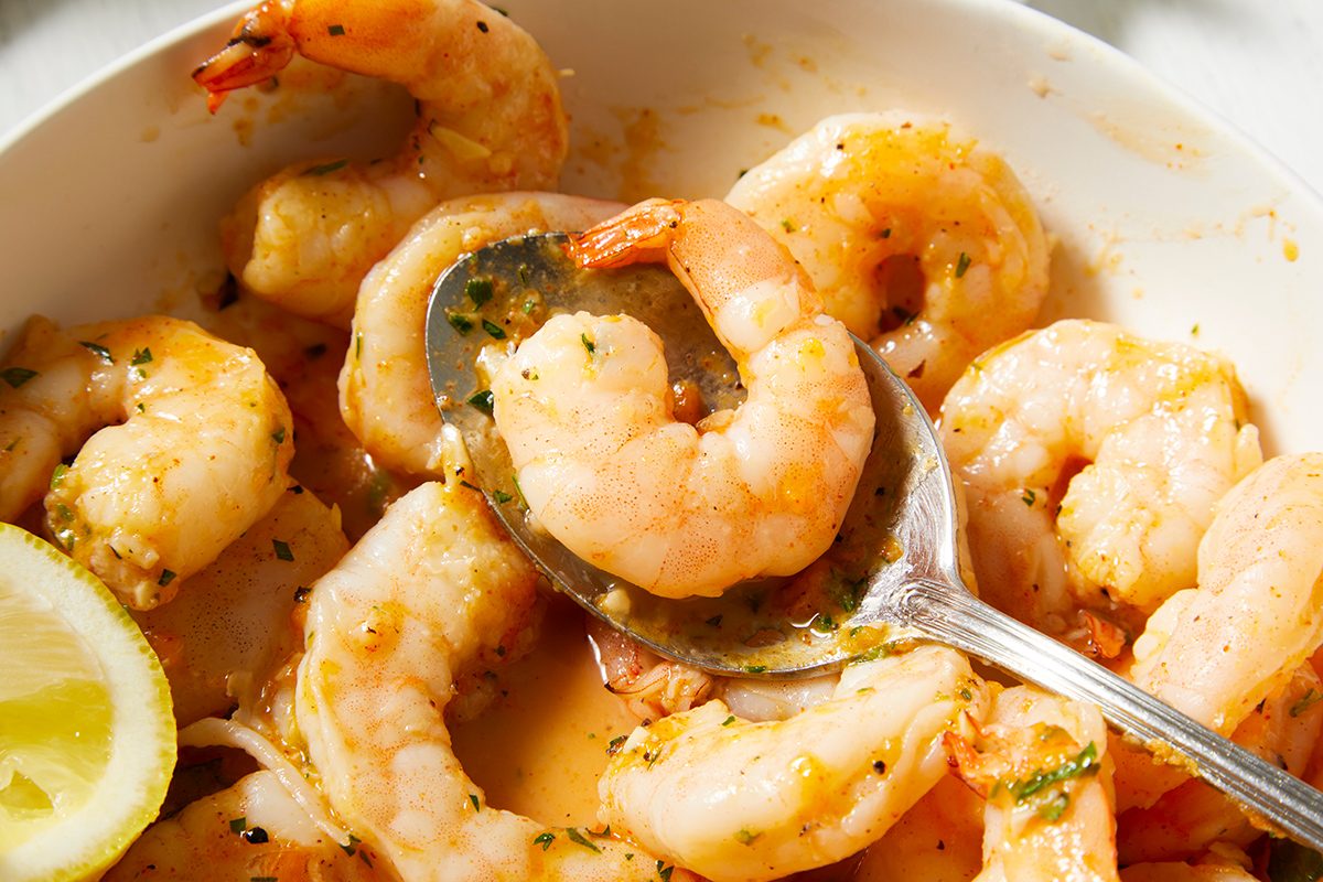 A bowl of cooked shrimp seasoned with herbs and spices, with a metal spoon resting among the shrimp. A lemon wedge is visible in the lower left corner of the bowl.