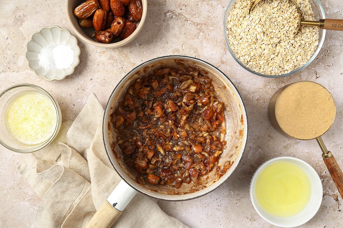 A saucepan filled with a cooked mixture of chopped dates and nuts is surrounded by bowls containing almonds, sesame seeds, a powder, a bowl of oil, melted butter, and a small dish of white salt on a light countertop.