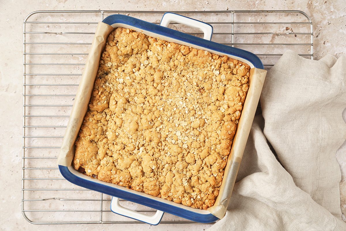 A square, blue baking dish filled with golden-brown crumble sits on a wire cooling rack, partially covered by a beige cloth. The crumble topping looks crisp and slightly uneven.