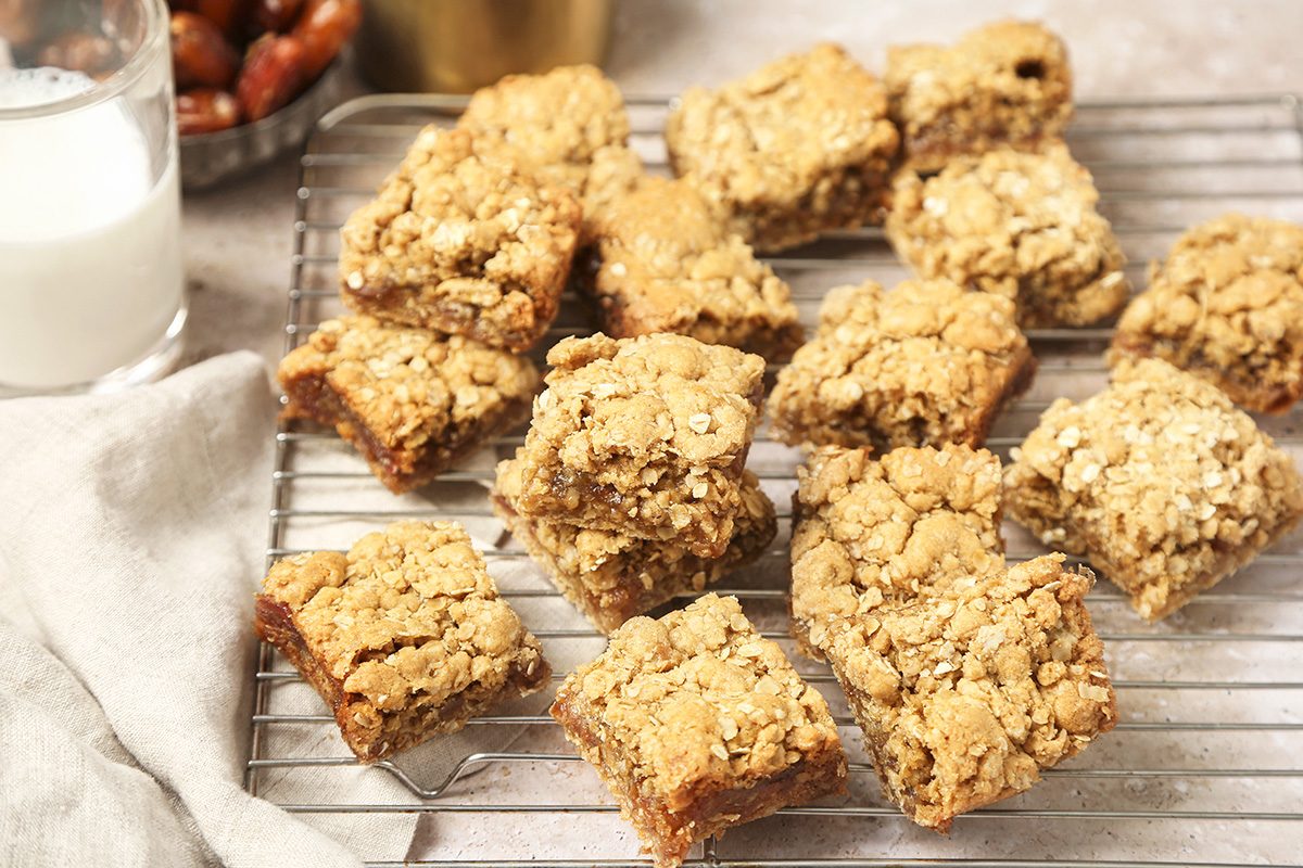 A cooling rack holds several oat and date bars with crumbly tops. A glass of milk and a bowl of dates are nearby on a light surface, with a light cloth partially visible on the side.