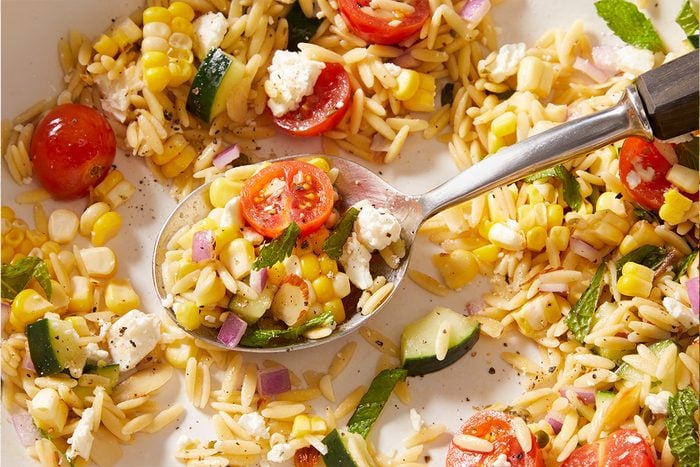 A close-up of a fresh orzo salad with cherry tomatoes, corn, cucumber, red onion, feta cheese, and herbs, served with a spoon.