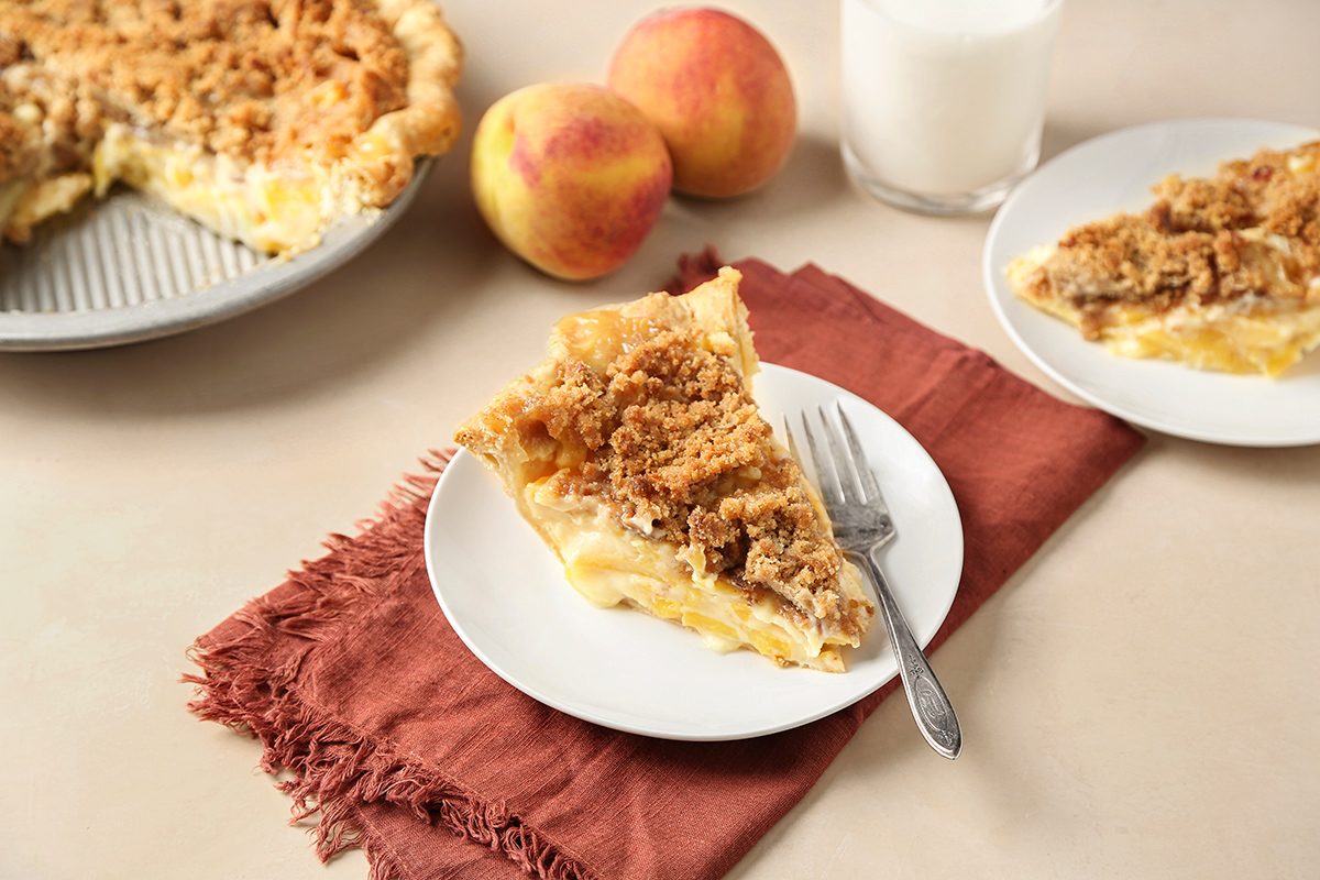 A slice of crumb-topped pie on a white plate with a fork rests on a brown napkin. Nearby are two peaches, a glass of milk, another slice of pie, and a partial pie in a tin.