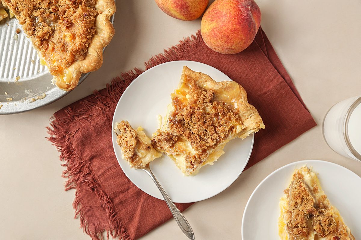 A slice of peach crumble pie on a white plate with a fork, next to a glass of milk, whole peaches, and a pie dish on a beige surface with a rust-colored napkin.