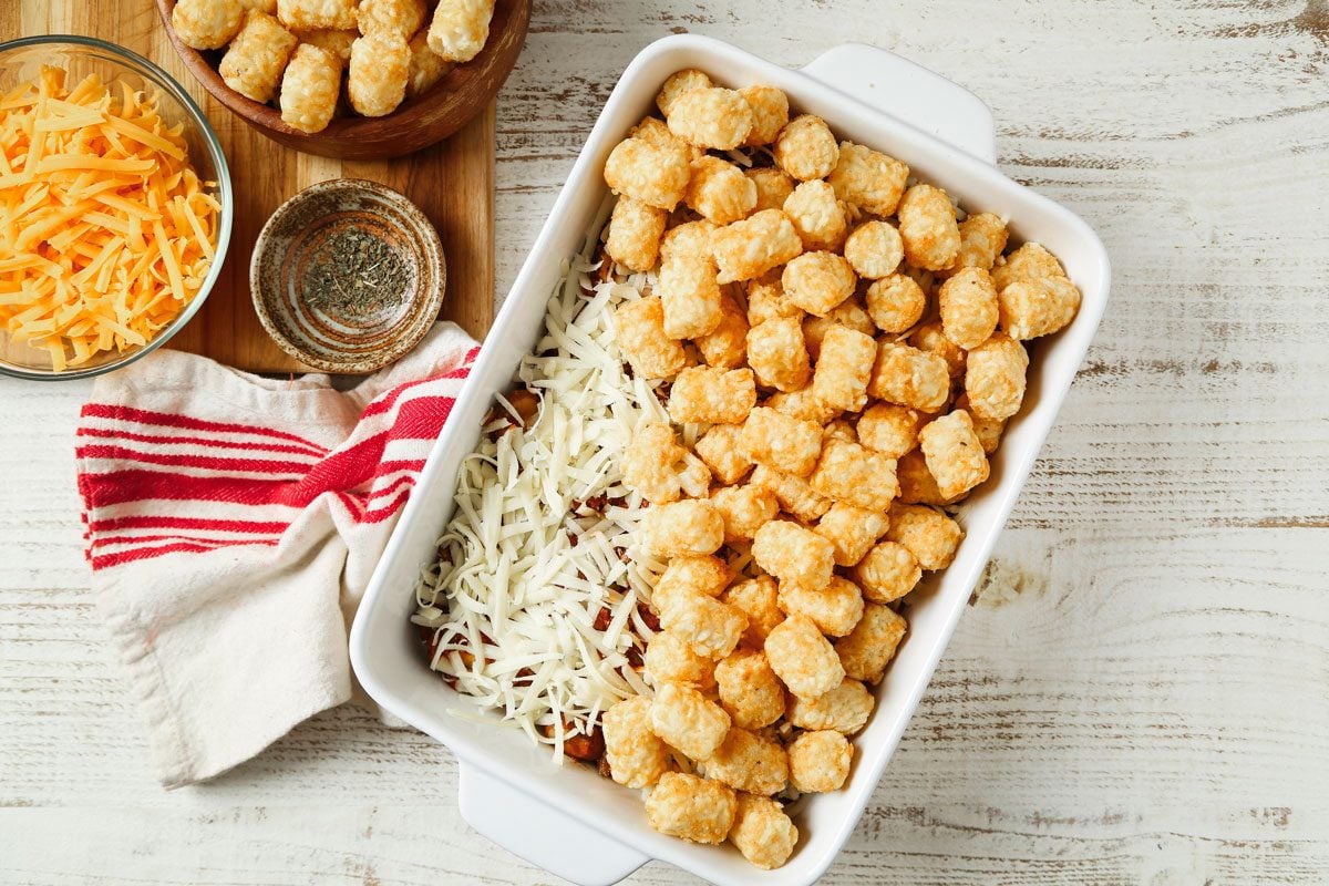 Overhead shot of transfer to a greased baking dish; Top with mozzarella cheese and potatoes; Bake uncovered at 400 degree until potatoes are lightly browned 30-35 minutes; cheese and potatoes bowls are on wooden cutting board; a napkin is nearby; all set on a wooden surface;