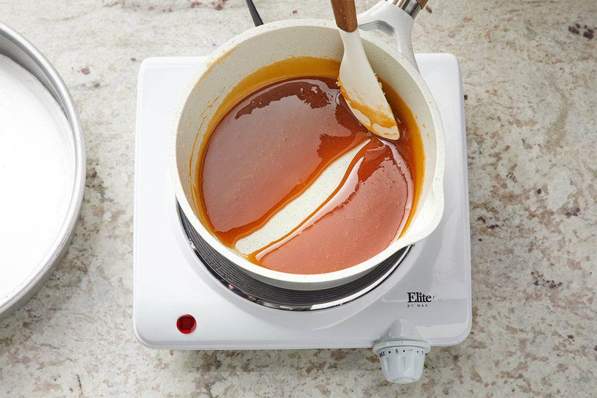 A pot of caramel sauce being stirred with a white spatula sits on a small electric stovetop, placed on a light-colored kitchen countertop.