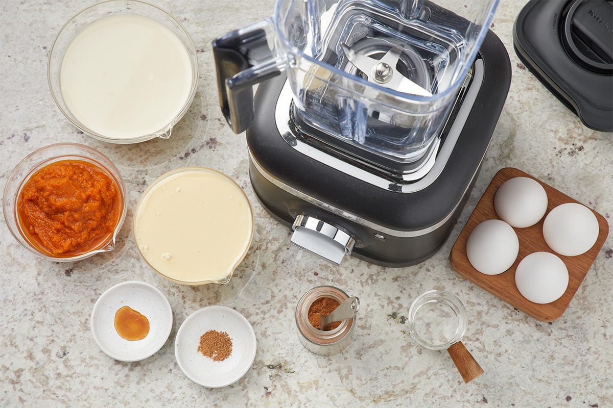 Overhead view of a blender surrounded by bowls of pumpkin puree, cream, sweetened condensed milk, spices, vanilla extract, four eggs in a holder, and a small jar, all on a light countertop.