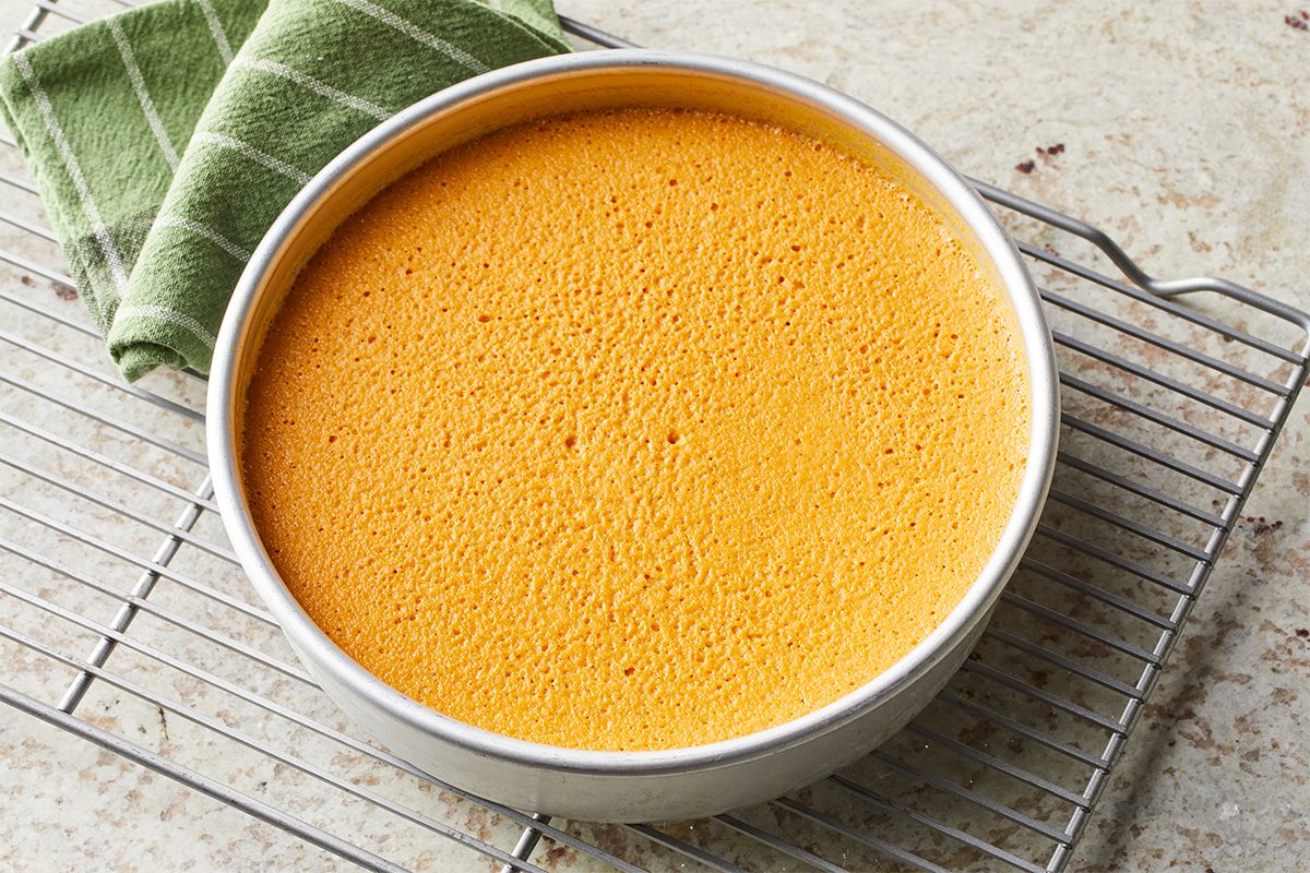 A round, golden-brown sponge cake in a metal pan cools on a wire rack, with a green striped kitchen towel nearby on a light countertop.