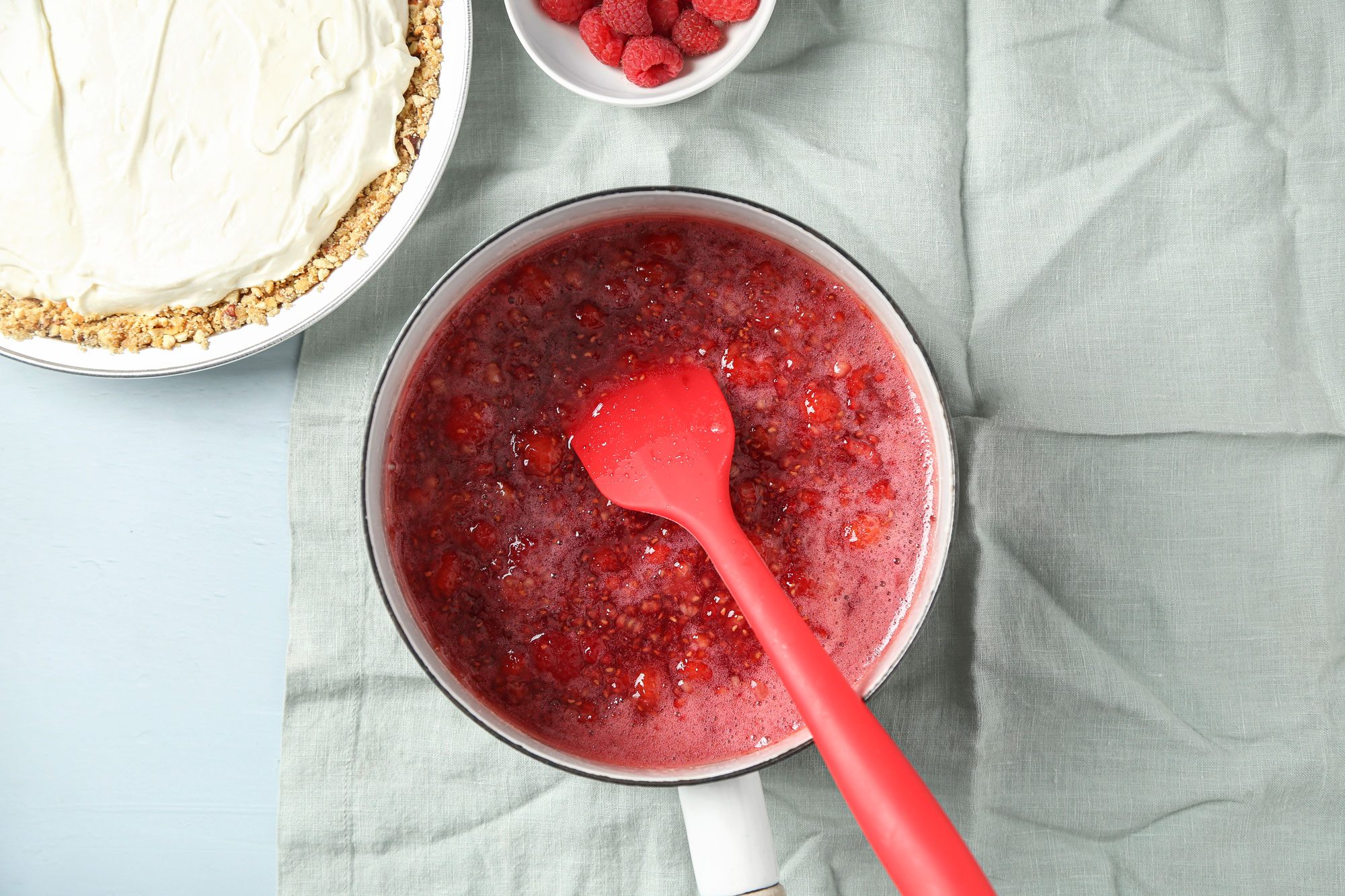 overhead shot of a saucepan with raspberry sauce and a red spatula sits on a light green cloth, Next to it are a bowl of fresh raspberries and a partially frosted dessert with a crumb crust