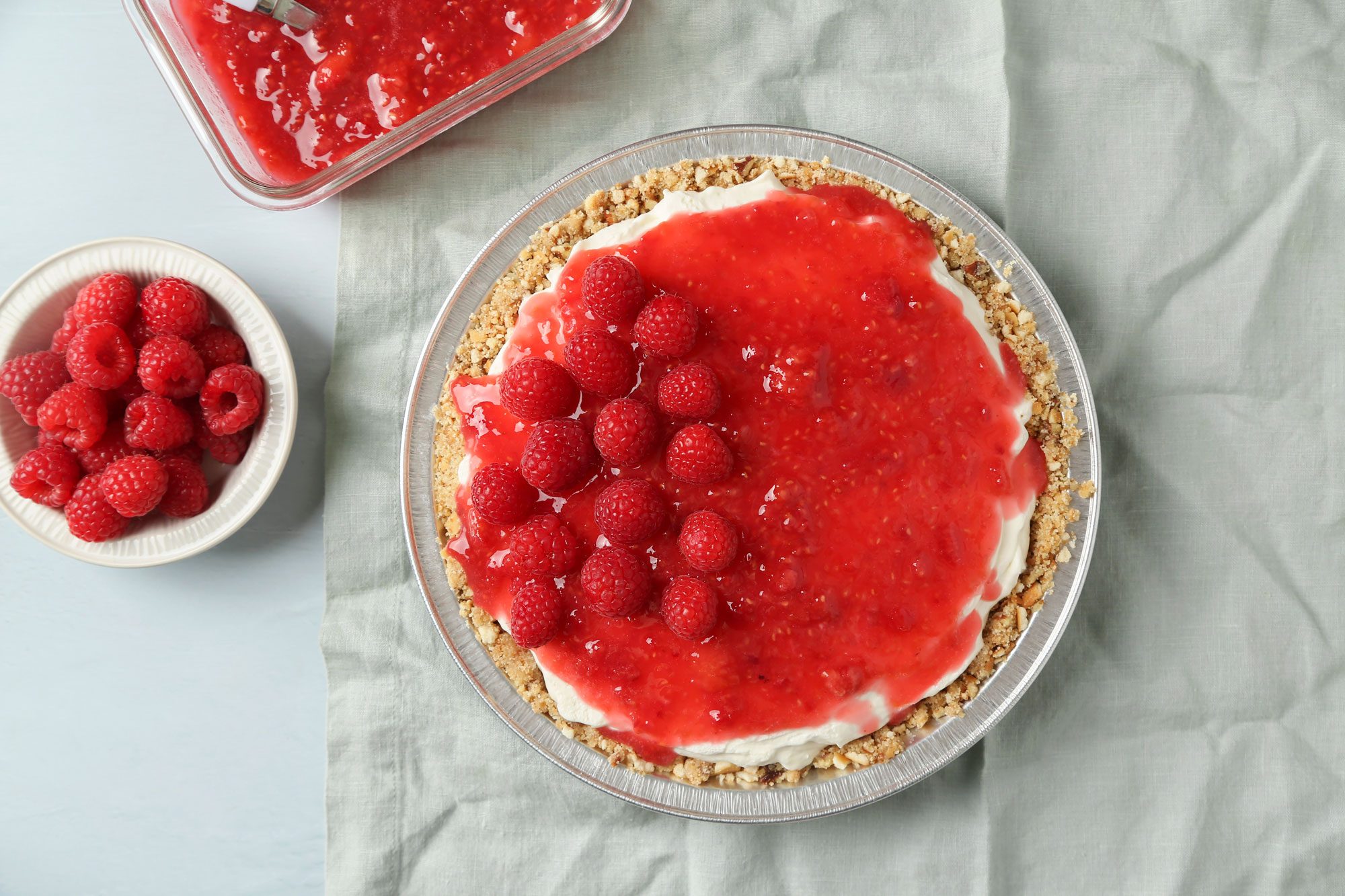 overhead shot of a cheesecake topped with fresh raspberries and raspberry sauce sits on a light cloth; a bowl of raspberries and a dish of extra sauce are nearby