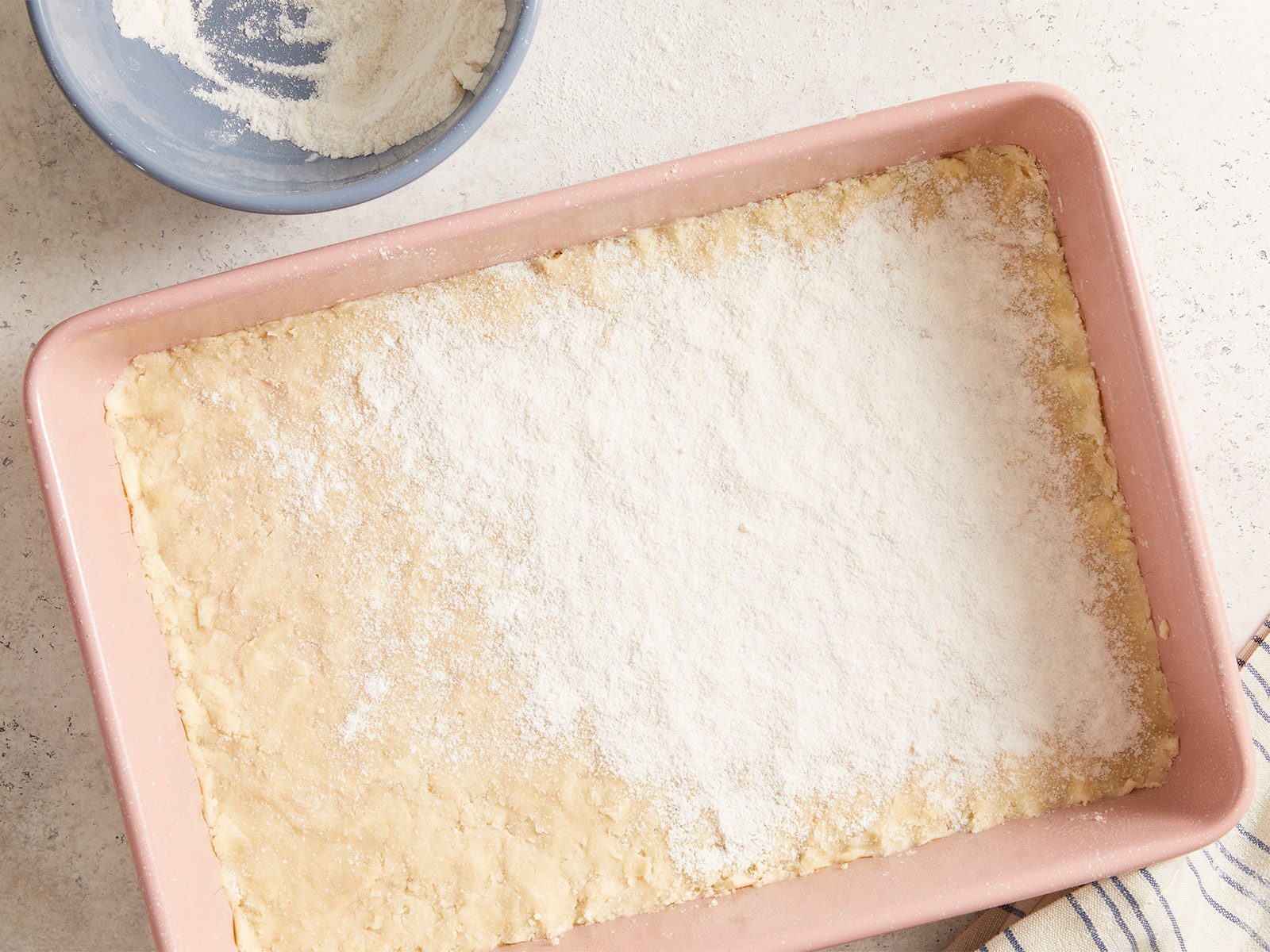 A pink baking dish filled with an unbaked dough, partially topped with a dusting of flour, sits on a light countertop. Nearby, a blue bowl with flour and a striped towel are visible.