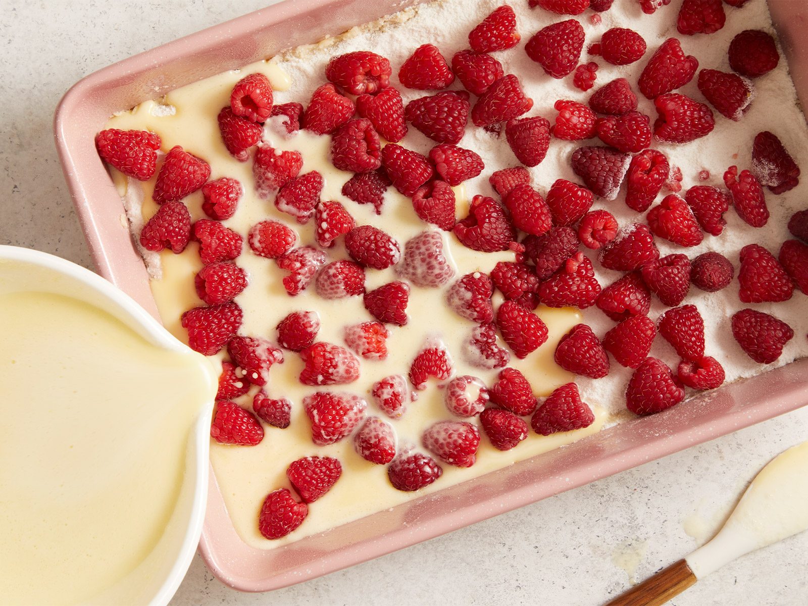 A baking dish filled with raspberries on a layer of batter, with creamy batter being poured over the raspberries from a bowl in the corner. A pastry brush lies nearby on the countertop.