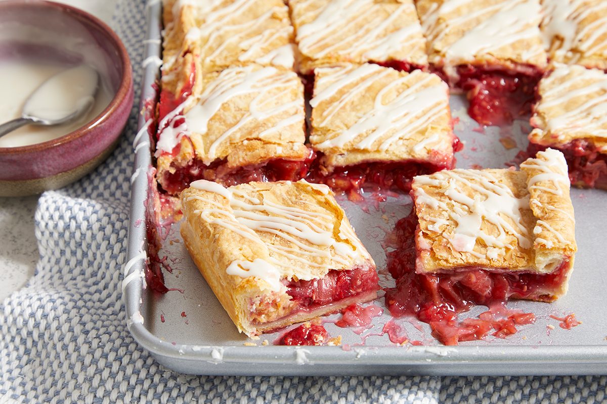 A metal baking tray with square slices of cherry-filled pastry, drizzled with white icing. A few pieces are separated from the rest, and there is a small bowl of extra icing nearby.