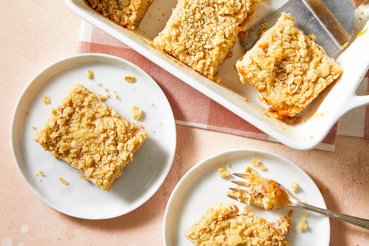 A baking dish with rectangular pieces of crumb-topped dessert, likely coffee cake or crumb bars. Two pieces are served on white plates, one with a fork, on a light surface with a checkered cloth underneath.