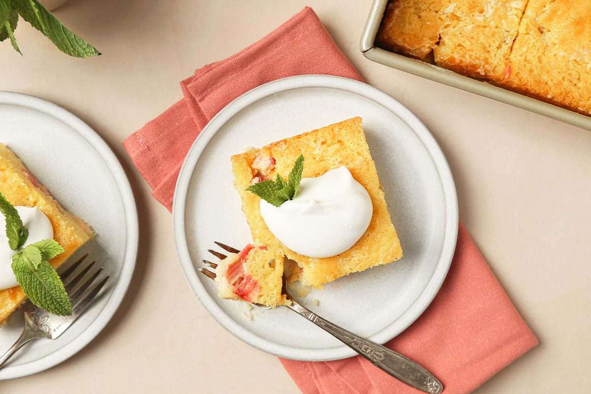 A slice of yellow cake topped with whipped cream and a mint leaf, served on a white plate with a fork. The plate is on a salmon-colored napkin next to a baking pan with more cake.