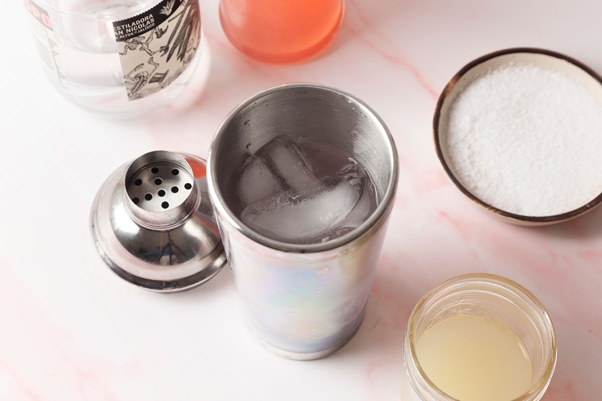 A metal cocktail shaker with ice sits on a pink surface, surrounded by bottles, a bowl of sugar, and a jar containing a light-colored liquid.