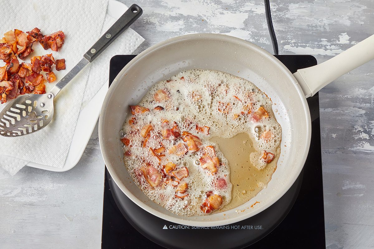 Chopped bacon pieces are sizzling and cooking in a white frying pan on a stovetop. A slotted spoon and cooked bacon rest on a paper towel nearby.