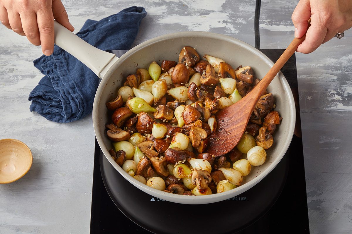 A person stirs a pan filled with pearl onions and mushrooms on an electric stovetop, using a wooden spoon. A blue cloth is nearby on the light gray countertop.