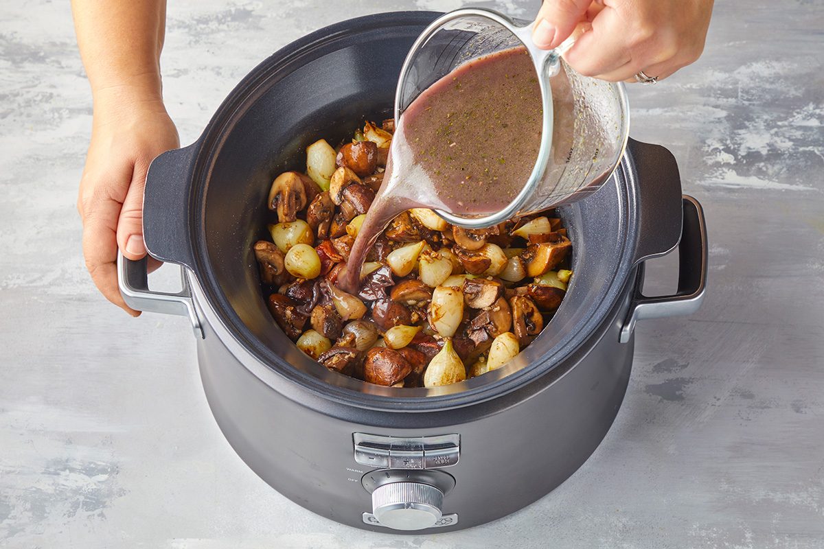 A person pours a dark liquid from a measuring cup into a slow cooker filled with potatoes, onions, and mushrooms.