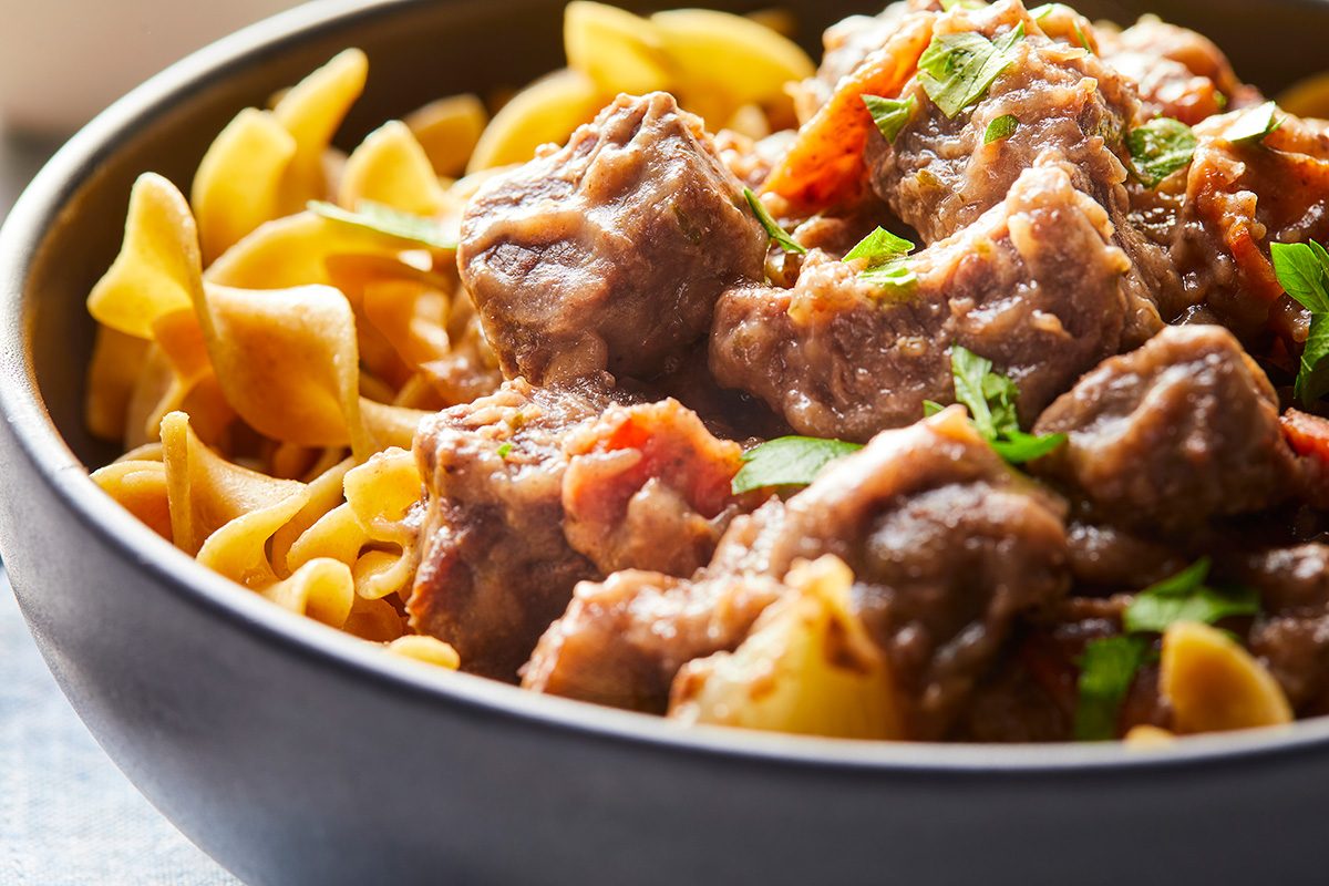 A close-up of a bowl filled with beef stew served over egg noodles, garnished with fresh chopped herbs. The beef looks tender, and the dish includes pieces of cooked vegetables.