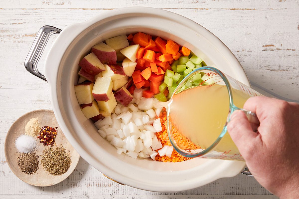 A hand pours broth from a measuring cup into a slow cooker filled with chopped potatoes, carrots, celery, onions, and red lentils. A plate with spices and seasonings sits beside the slow cooker on a white wooden surface.