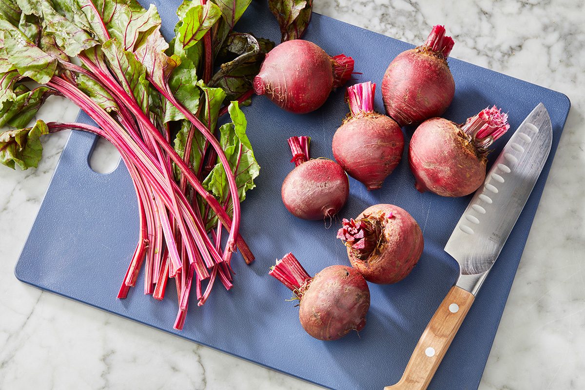 Whole beets with leaves and stems are on a blue cutting board next to a kitchen knife. The board is placed on a marble surface. Some beets are partially trimmed and some stems are separated.