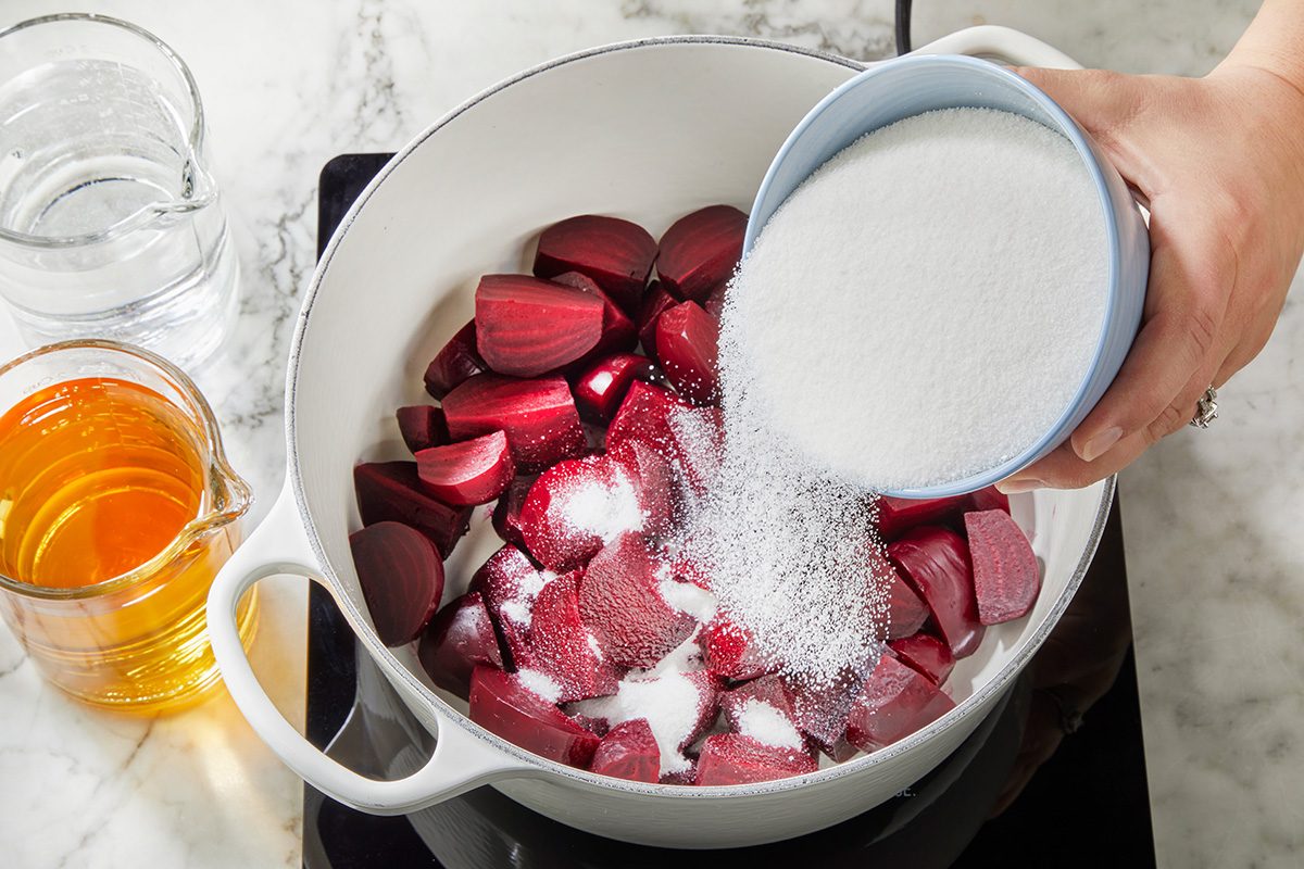 A hand pours granulated sugar from a cup into a white pot filled with chopped rhubarb. A glass of water and a glass of honey sit nearby on a marble countertop.