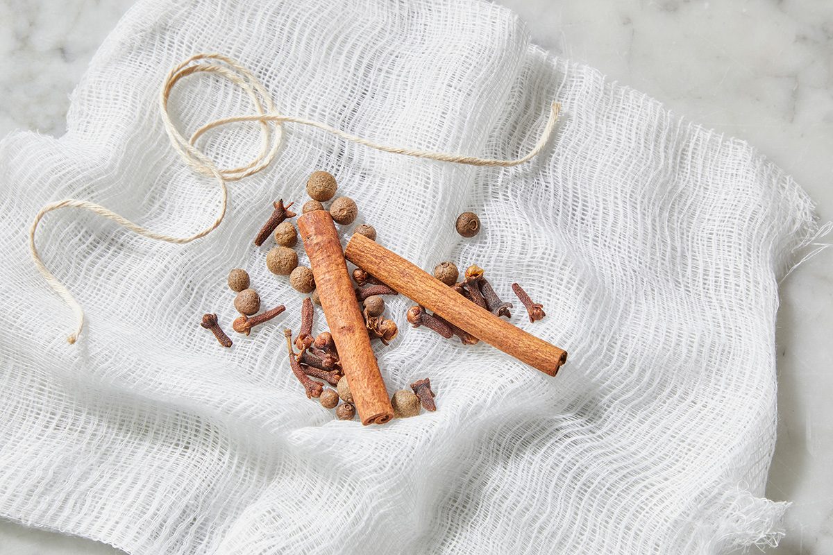 Two cinnamon sticks, whole cloves, allspice berries, and a piece of string are placed on a piece of white cheesecloth, set on a marble surface.