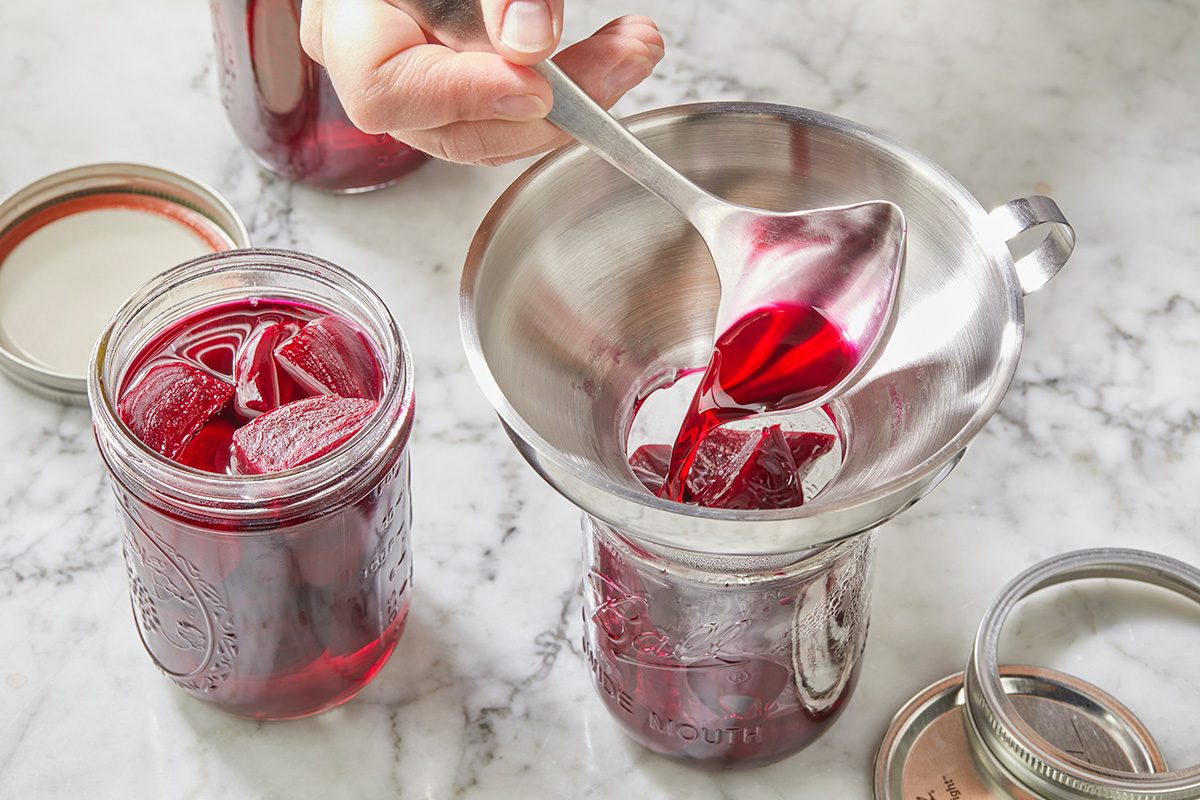 A hand pours red liquid from a ladle through a funnel into a glass jar on a marble surface; a filled jar with pieces of fruit and open jar lids are nearby.