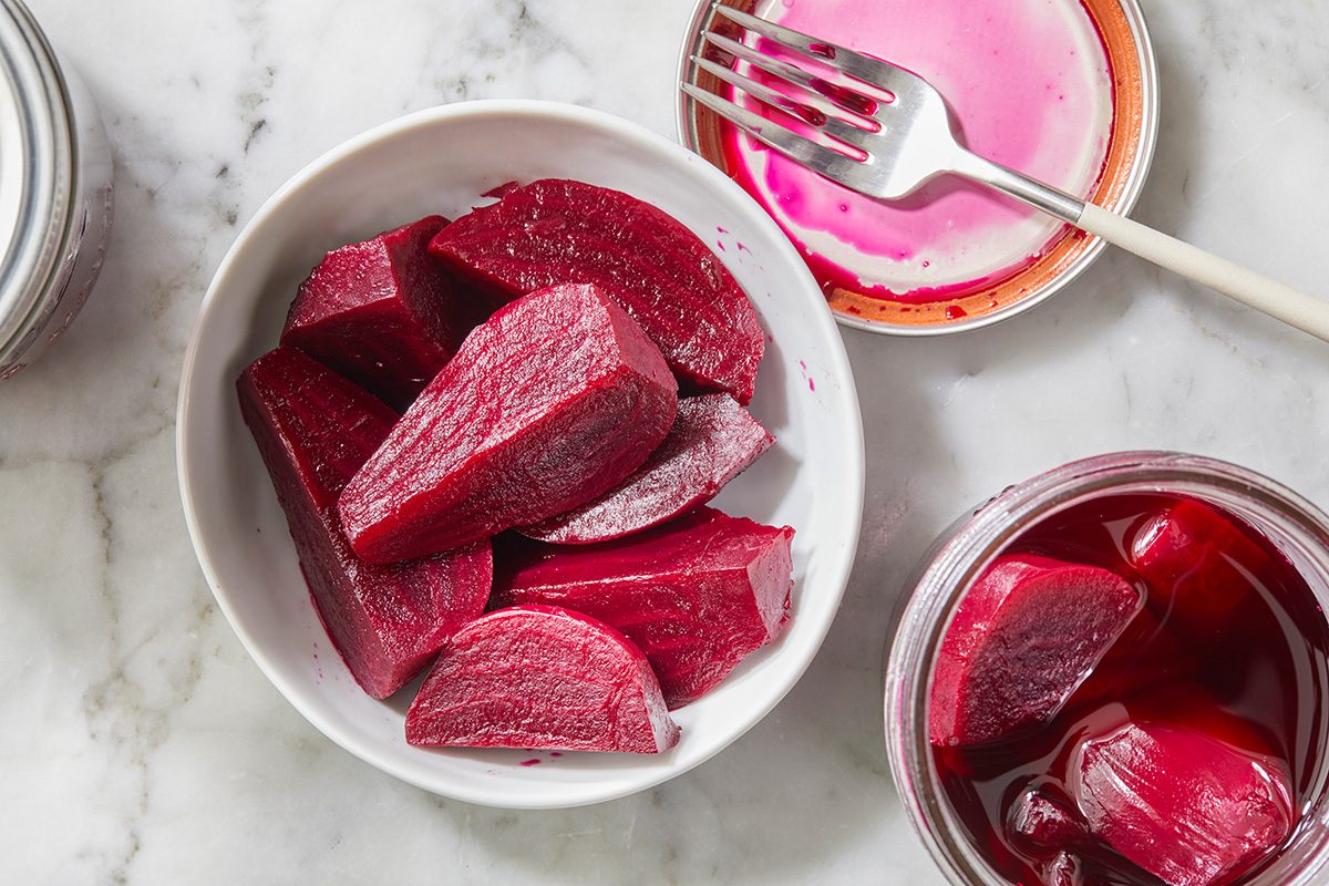 A white bowl filled with sliced pickled beets sits on a marble surface, next to a fork with pink beet juice and an open jar containing more pickled beets.