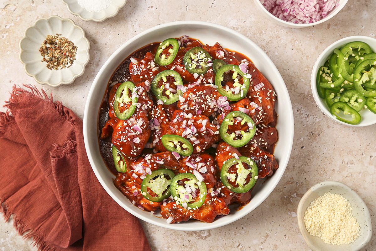 overhead shot of a bowl of spicy chili garnished with sliced jalapeños, chopped red onions, sesame seeds, and cheese, surrounded by small dishes of seasoning, grated cheese, and a red cloth on a light colored surface