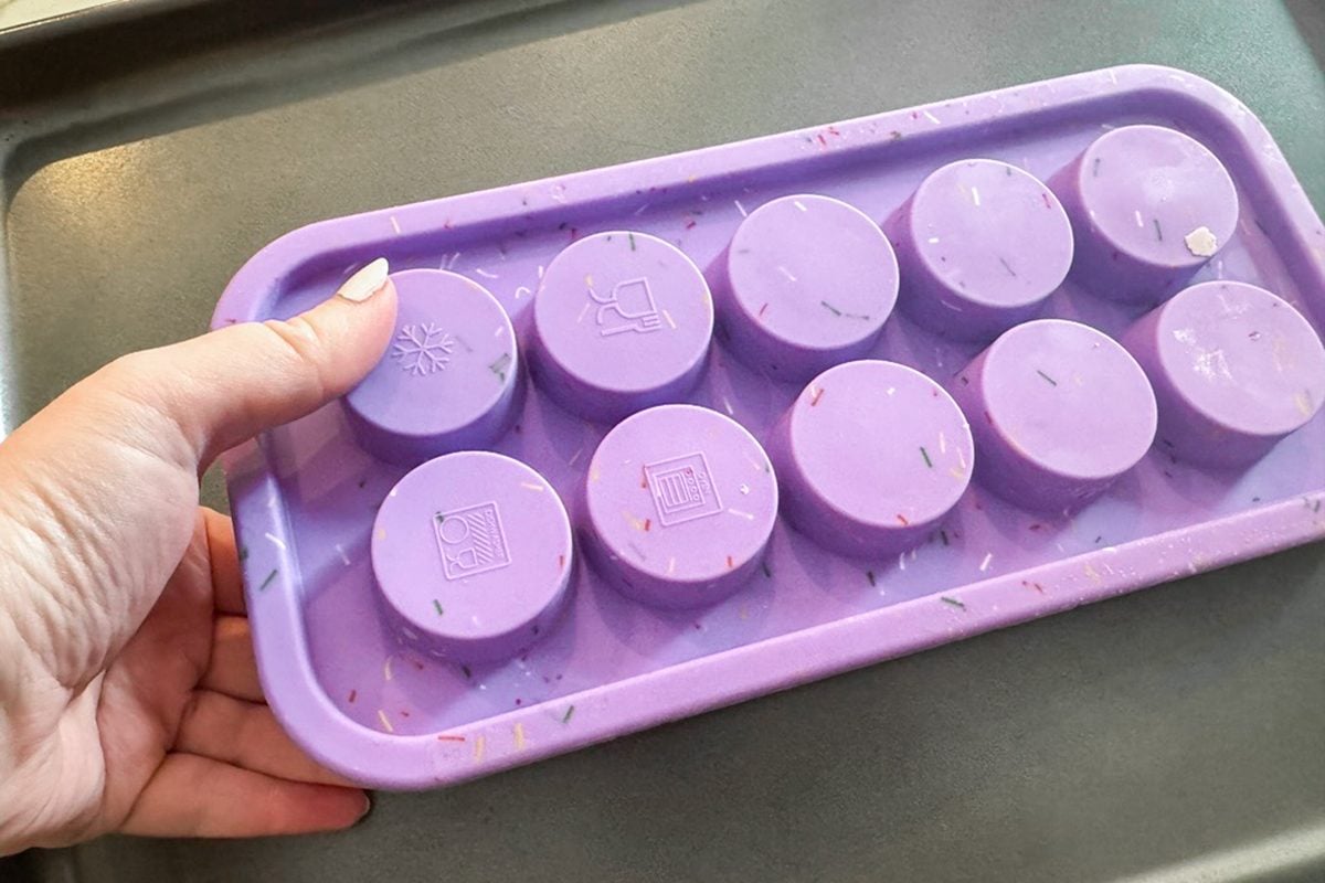 A hand holds a purple silicone mold tray with ten round compartments, each featuring a different symbol, over a gray surface.