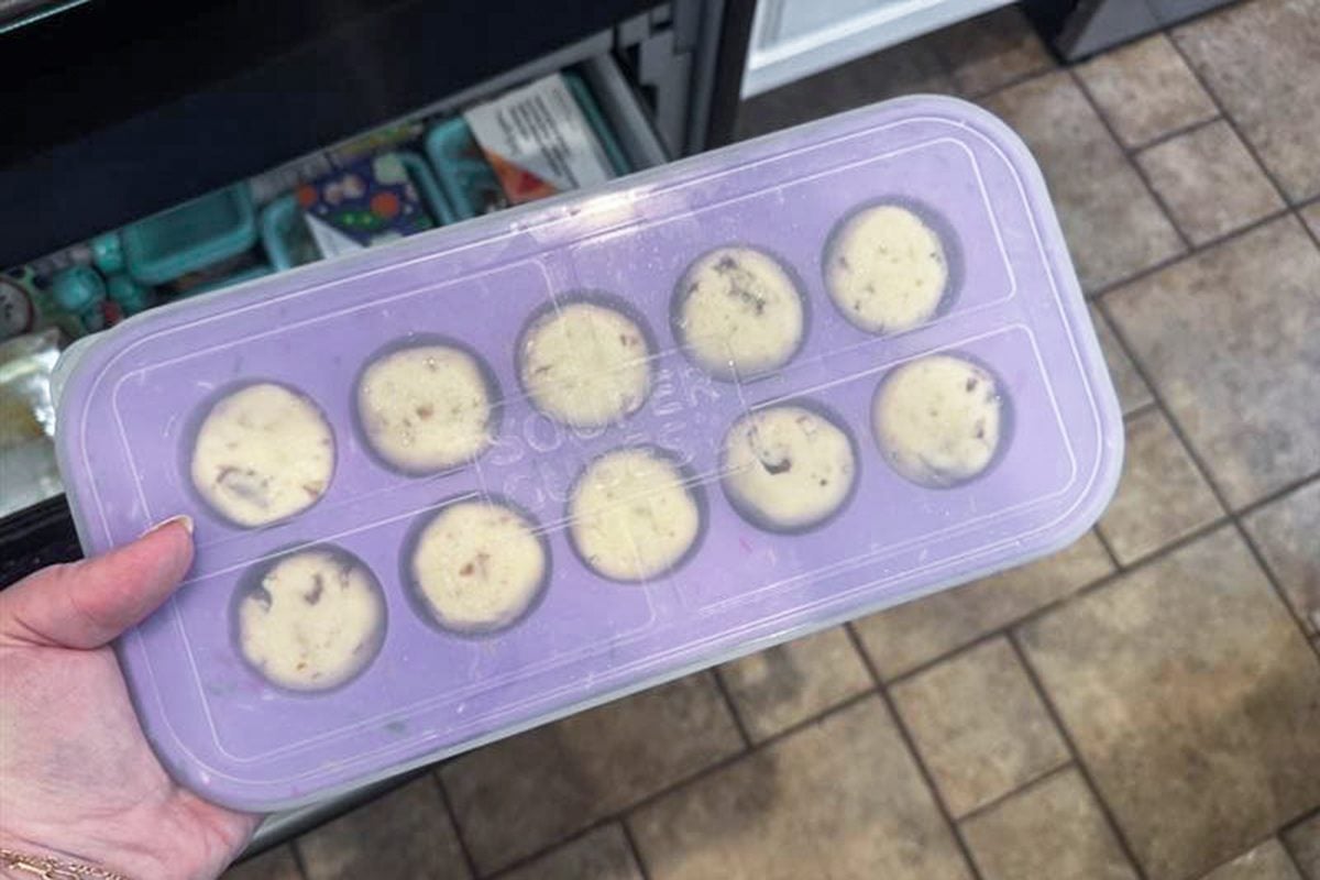 A hand holds a purple silicone tray with ten round compartments, each filled with cookie dough, above a tiled kitchen floor near an open freezer drawer.