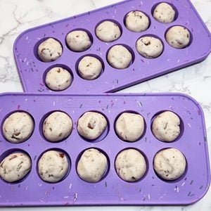 Two purple silicone baking molds, each holding ten unbaked, round cookie dough balls with chocolate chips, are placed on a white marble surface.